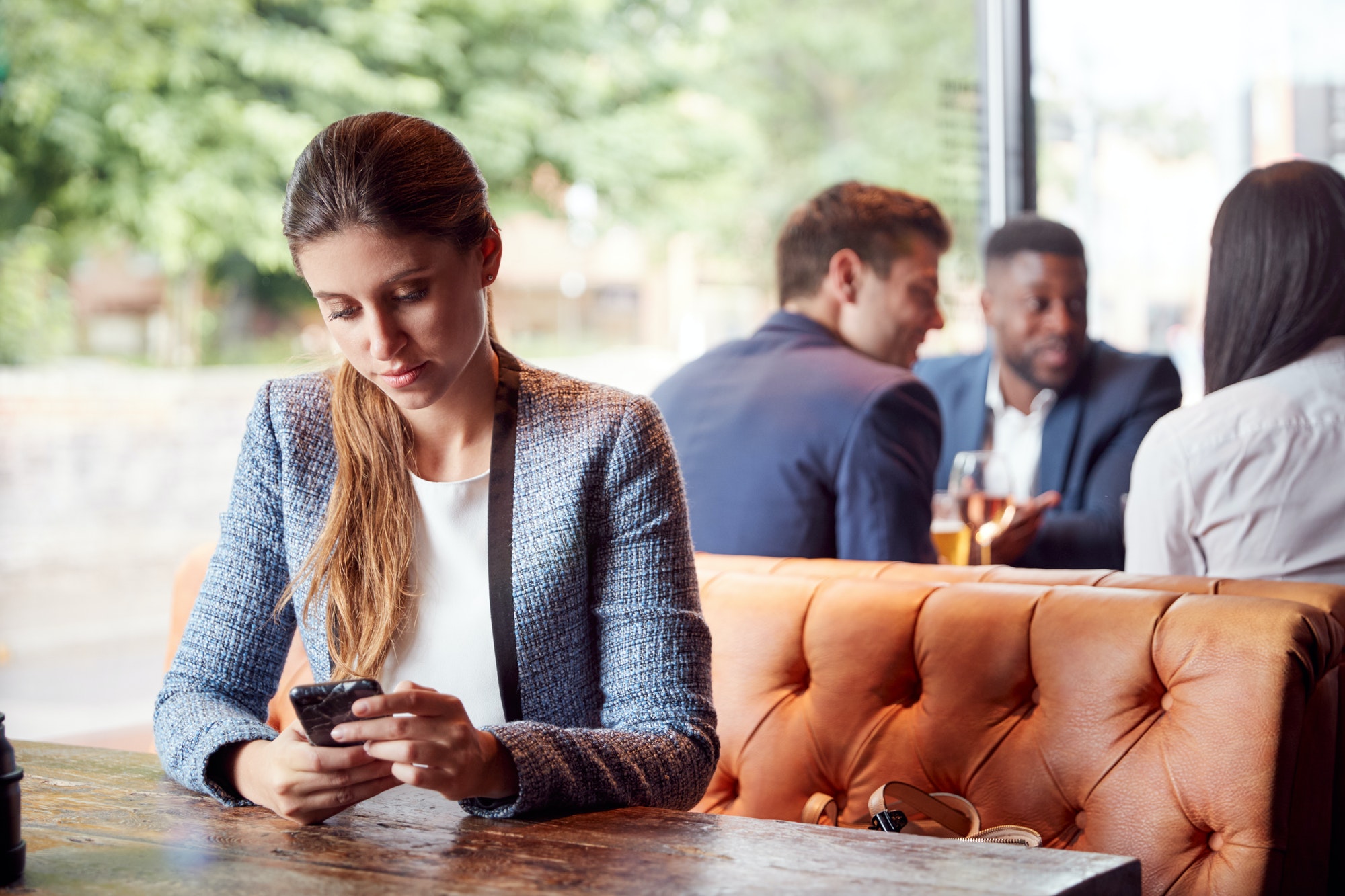 Businesswoman Sitting In Bar Checking Mobile Phones Whilst Colleagues Meet For Drink In Background